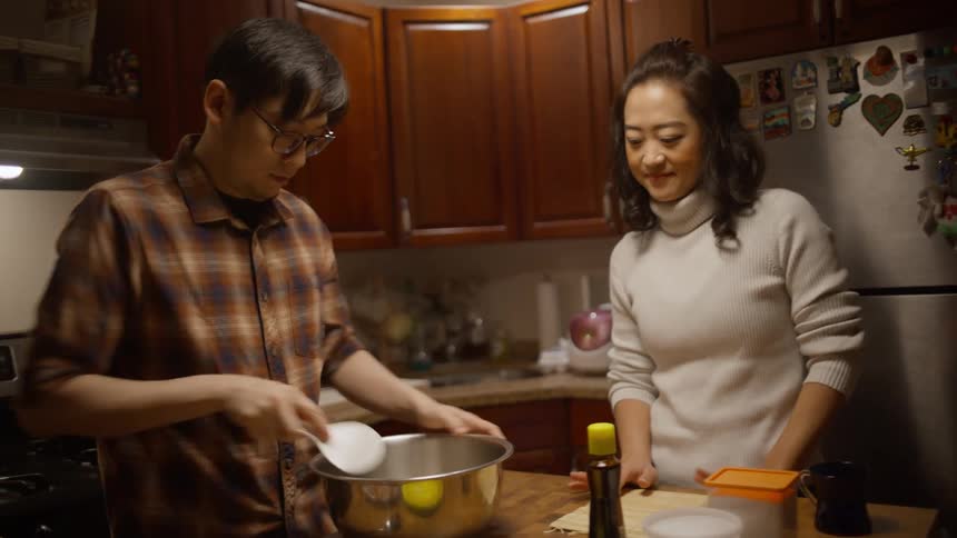 A man and a woman pour sauce into a mixing bowl in a kitchen