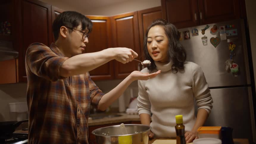Man cooking in the kitchen and offering a spoon to his wife for tasting who approves