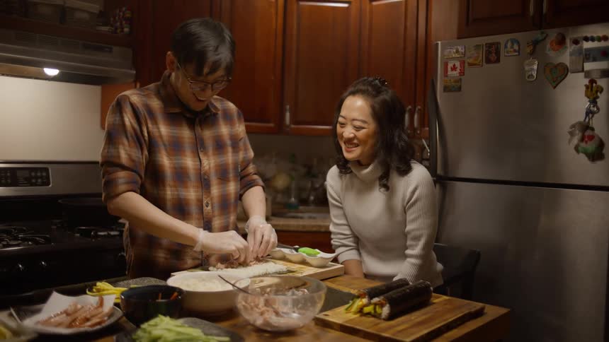 A couple smiling and laughing in a kitchen as the man prepares sushi rolls