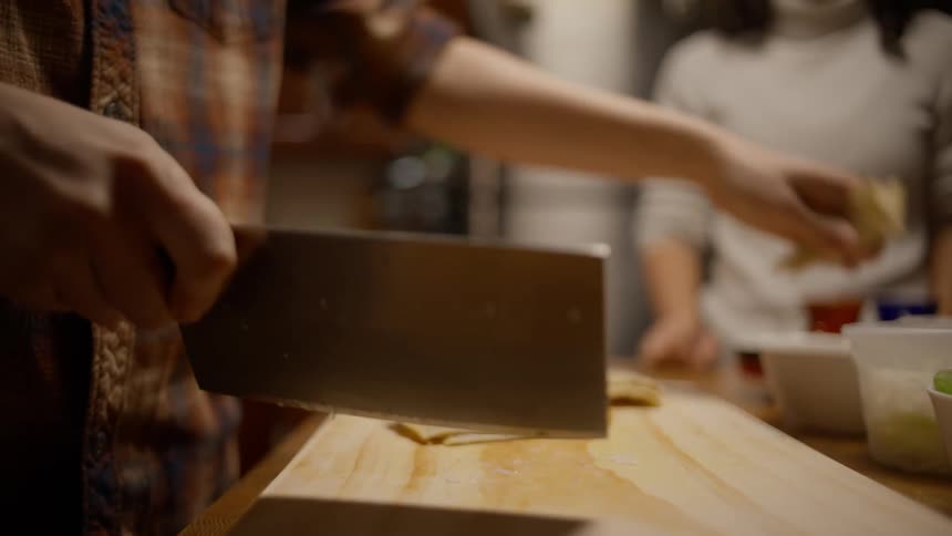 A person using a cleaver is cutting vegetables on a wooden board