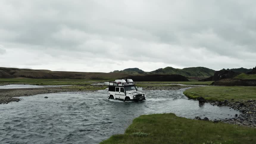 White jeep with gear strapped on top driving across a water covered road section surrounded by mossy green flat land and hills with mountain peaks and a white clouded sky in the background