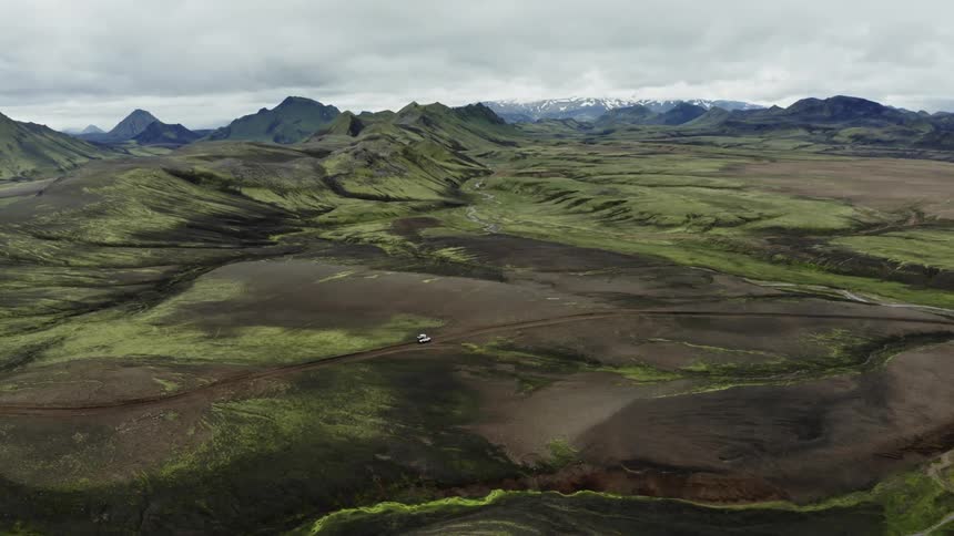 Aerial of white jeep on a single path driving across rocky green highland with mountains and clouded sky
