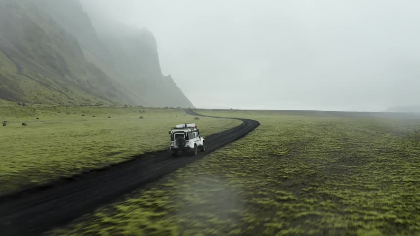 An aerial view of a sport utility vehicle traveling along a dirt road on a large plain next to a mountain on a damp foggy day