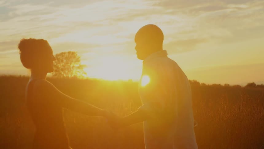 A man and woman hold hands and walk in the grass as the sunsets
