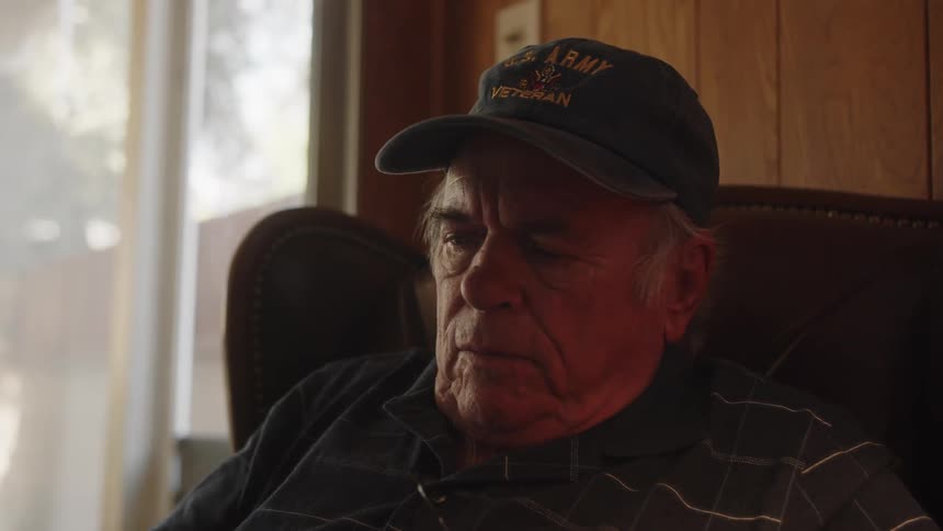 Elderly man wearing a blue cap and shirt sits in armchair laughing