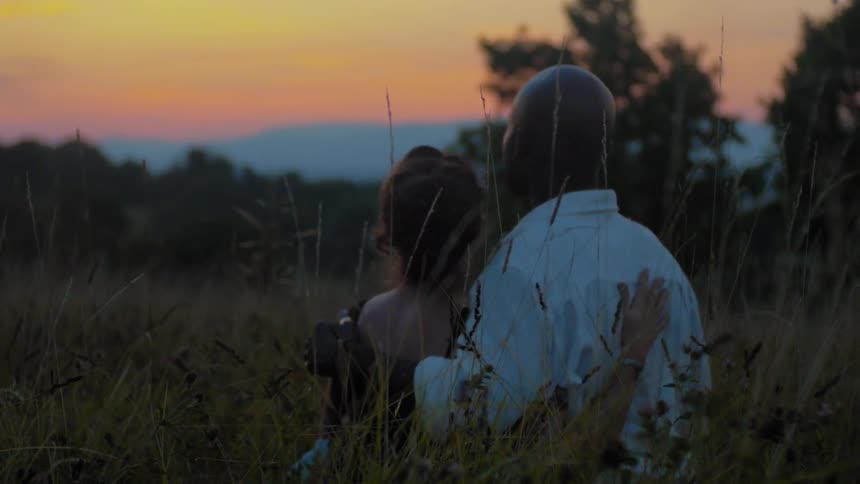 A man and woman embrace in the grass as they watch the sunset