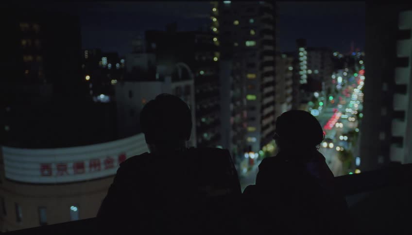 Friends on the balcony of a building chatting at night facing a bustling street with cars
