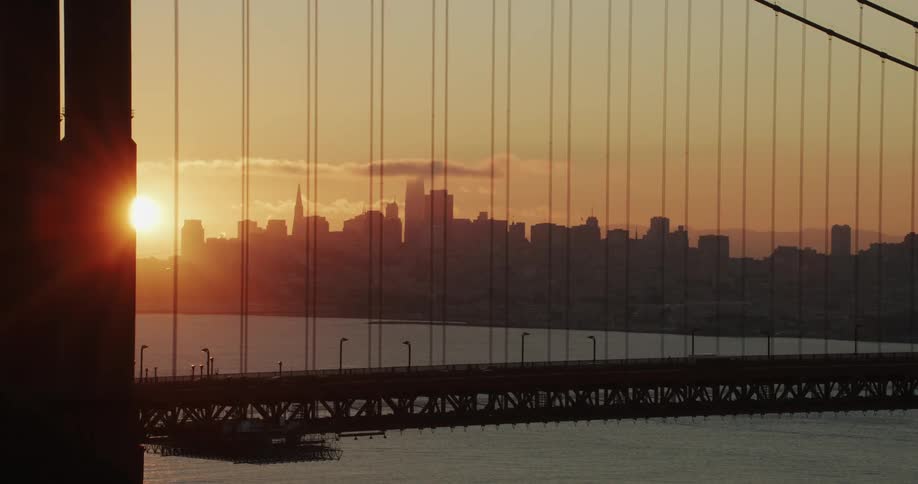 Traffic moves over a suspension bridge as the sun glows on the horizon behind the metropolitan city skyline
