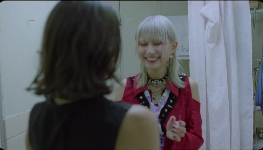 A woman exits the bathroom stall notices another woman applying makeup and politely requests to borrow the eyeliner