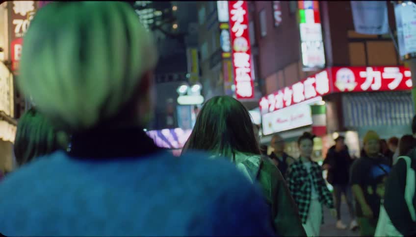 Several people walking on a sidewalk and there are many lights on commercial billboards