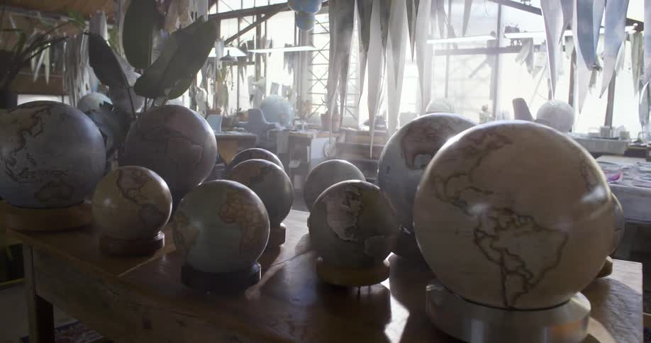 White globes with black continent outlines in different sizes sit on a display table in a workroom with other workstations and sunlight coming in through windows in the background