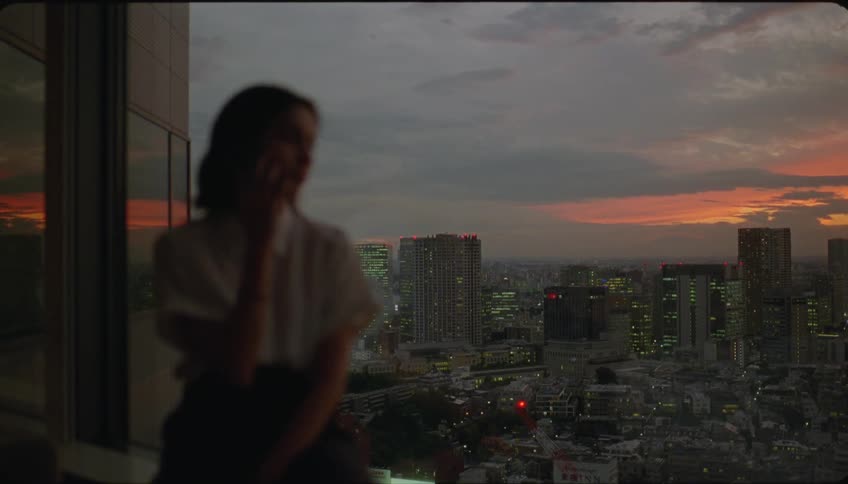 Woman on an apartment balcony talking on the phone looking at the city view during sunrise