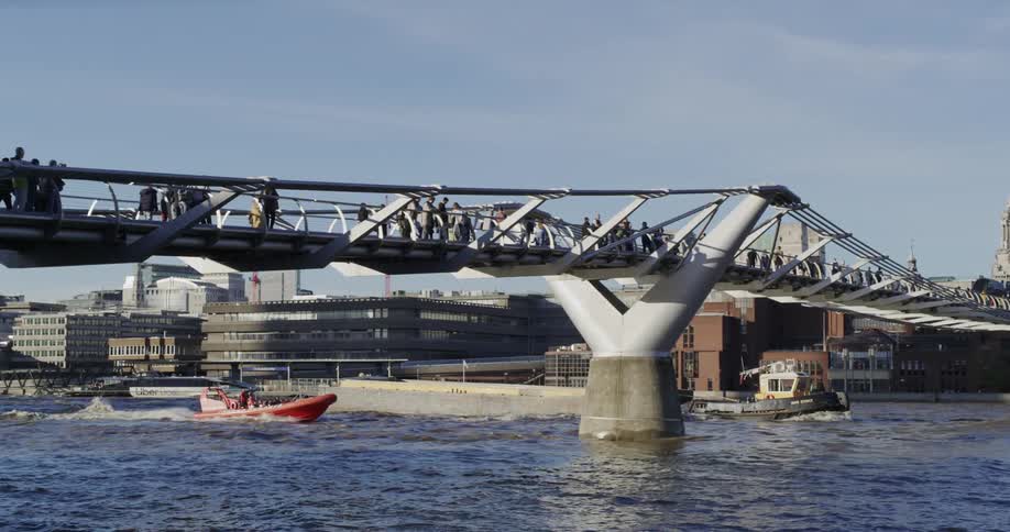 People walking on a bridge over the sea with tourist boats passing just below