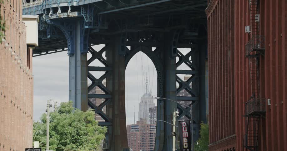 Empire state building in new york under a hazy pale sky viewed through an arch under a large tall bridge