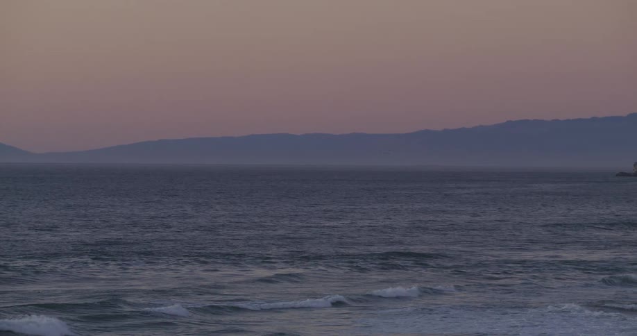 Waves roll into shore with mountains in the distance under a hazy sky and a small building on top of a promontory along the coast