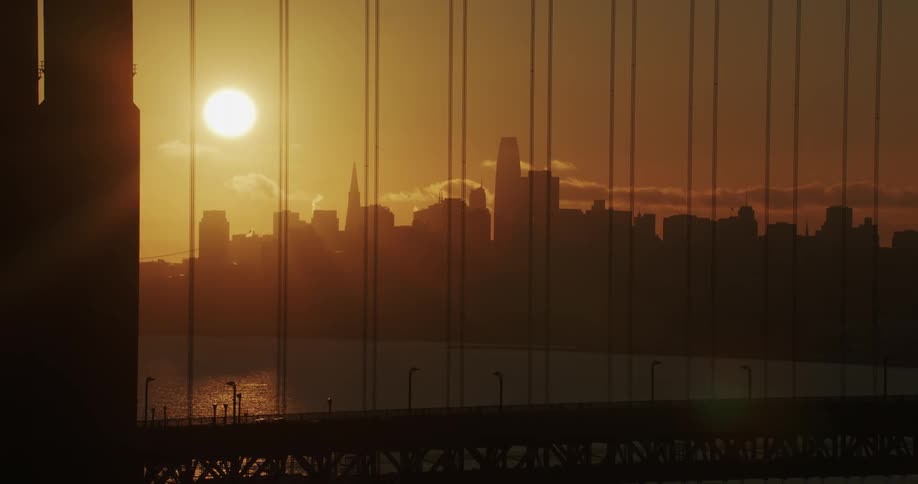 View through a suspension bridge of the sun glowing on the horizon behind the metropolitan city skyline