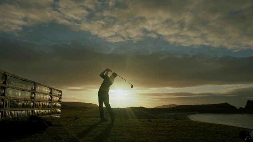 Silhouetted golfer taking a swing at the ball on a green golf course in the direction of the bright rising sun with clouded blue sky