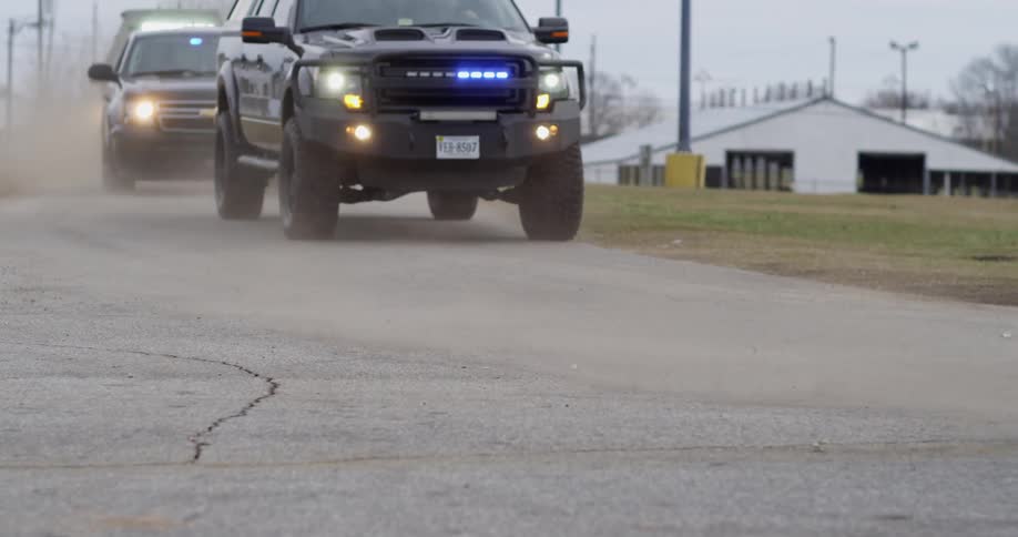 Unmarked police vehicles flash blue and red lights while driving along road past warehouses