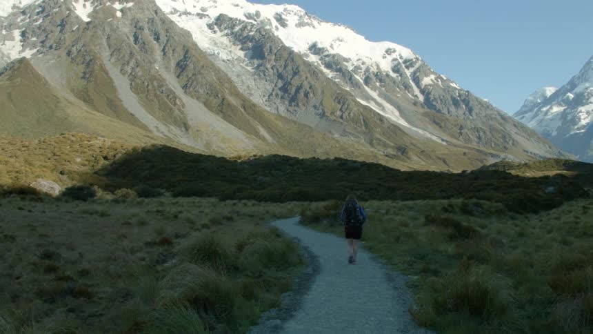 A person is hiking on a small trail towards a snow covered mountain