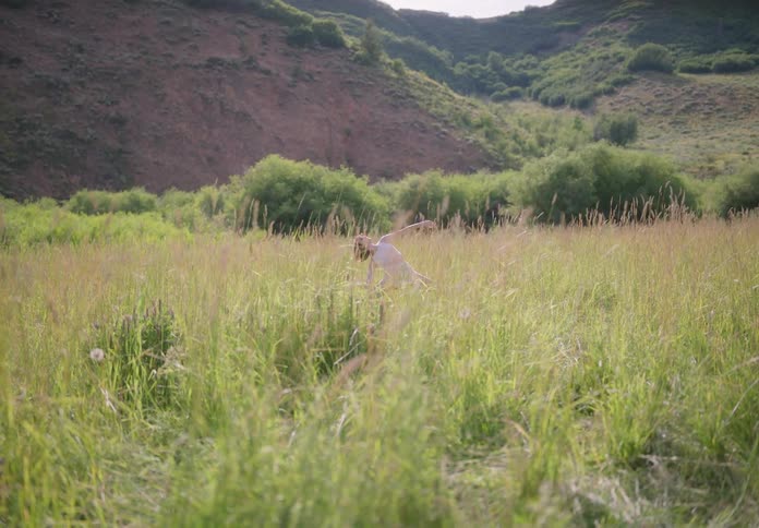 Woman in white dancing in the midst of tall grass in a grassland with mountains around in daylight