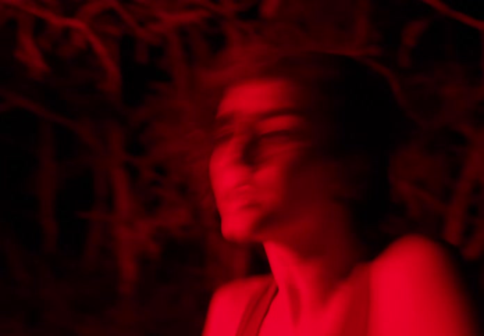 Portrait of a young woman moving her body chaotically with red light underneath her in a forest at night