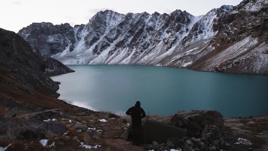 Panorama of a man facing a blue lake with snowcovered mountains
