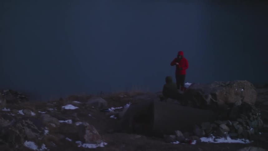 Two people on a rock facing a lake surrounded by snowcapped mountains