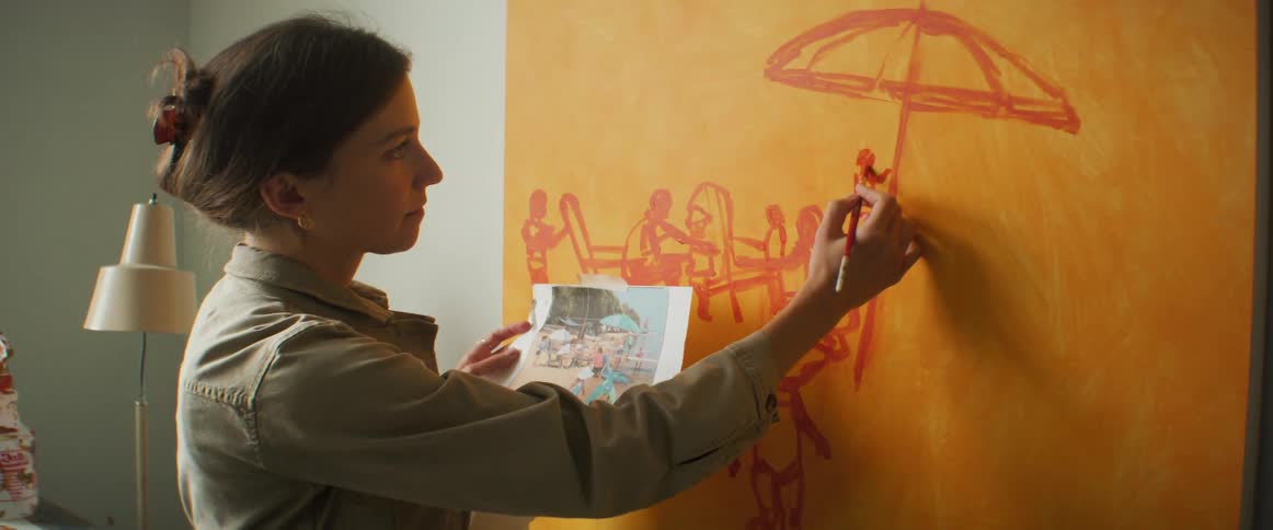 Painter painting on a canvas on the wall while holding a printed image in her other hand