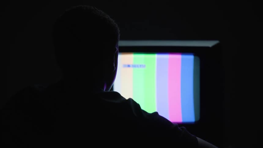 A man sitting in the dark staring at a tv displaying rainbow lines