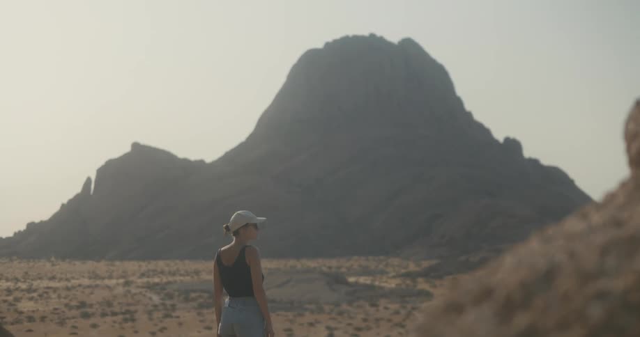 A woman dressed in a cap sunglasses dark top and gray pants observes a rocky mountain