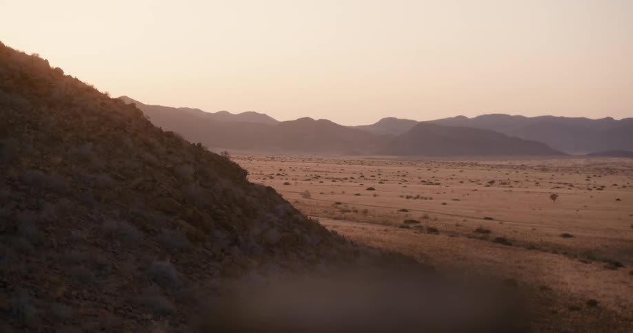 A desert landscape with a mountain slope in the foreground and a mountain range in the distant background