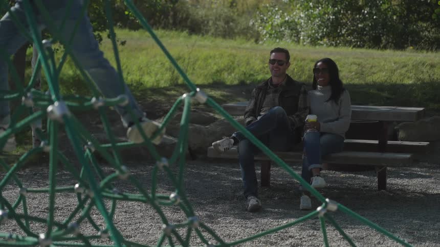 A couple siitting on a park bench watching their kid play as they smile