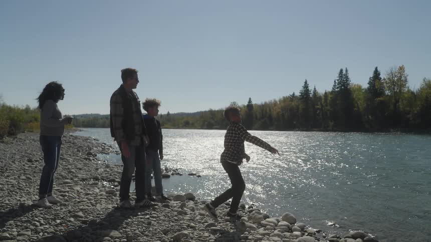 A boy throws stone on the river at daytime while his family stood nearby to watch