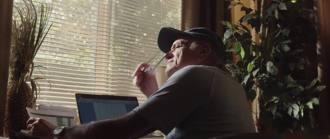An elderly man wearing a cap and a tshirt engrossed in thoughts works on a laptop