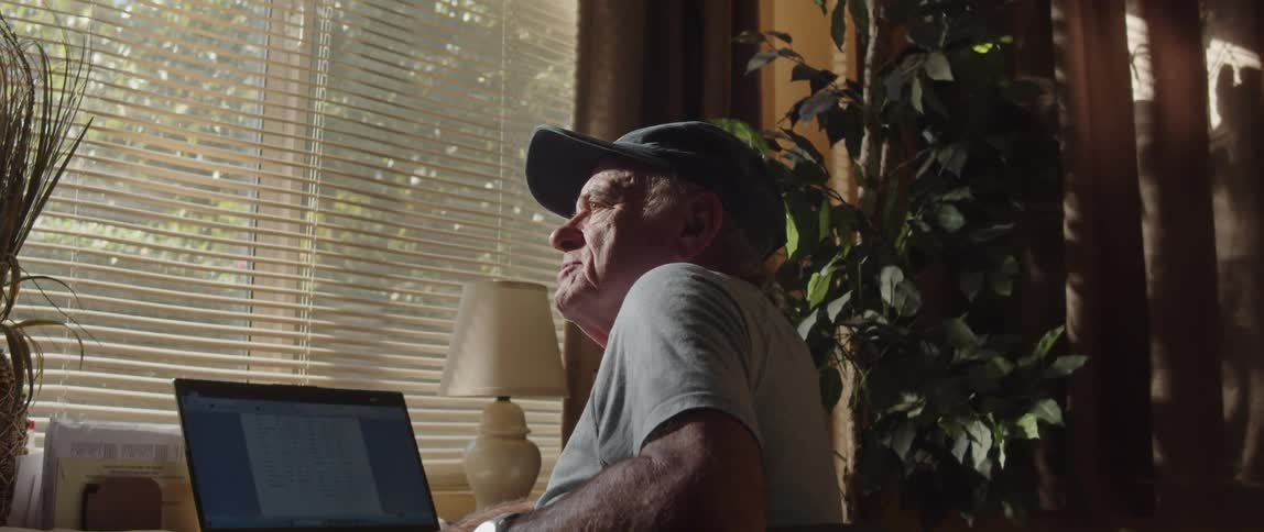 Elderly man sits in front of laptop and thinks while looking out window