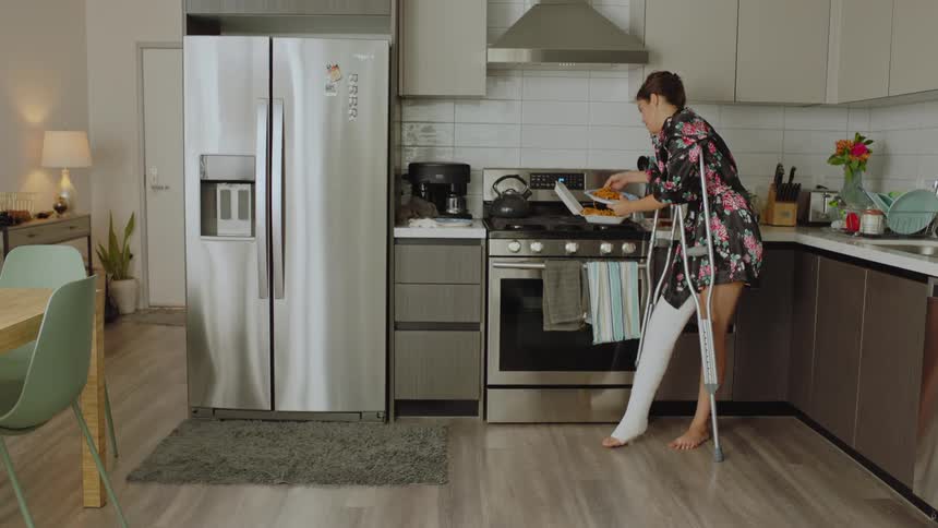 Woman on crutches putting pasta on her plate while talking then accidentally dropping the pasta on the kitchen floor