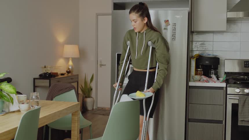 Woman on crutches prepares food in kitchen and hops to table to eat