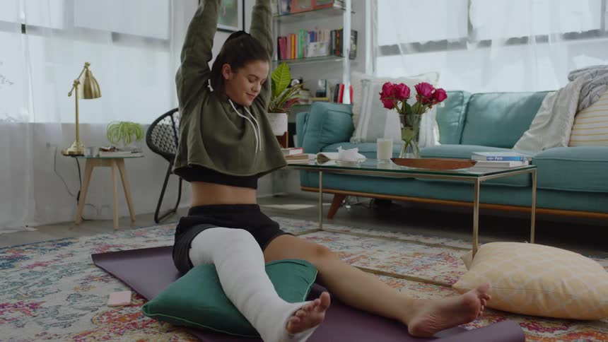 A woman is on a floor mat in her living room stretching while wearing a leg cast during the day