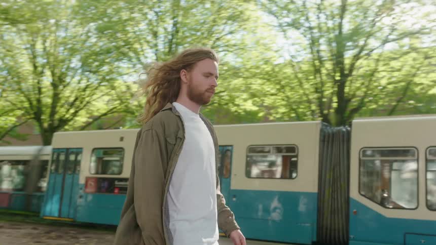 Young long haired man rides a skateboard on a road in the city following a train