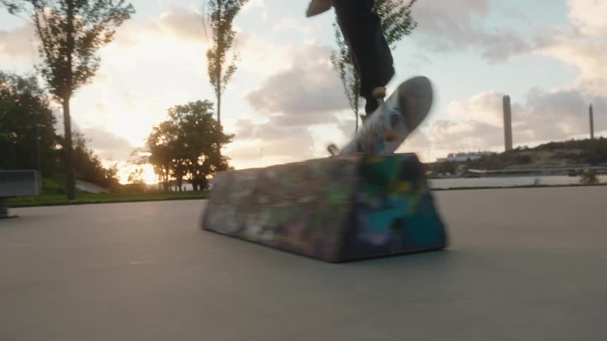 Male skateboarder with long curly hair jacket jeans and brown shoes skateboards on concrete road doing a hard flip at sunset