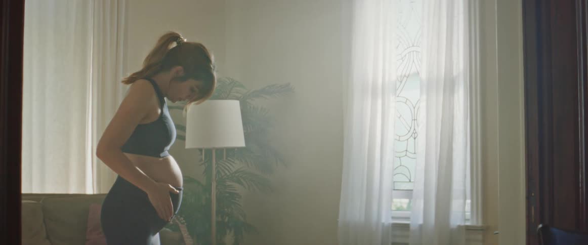 A pregnant woman in a sports bra and leggings stretches in her living room