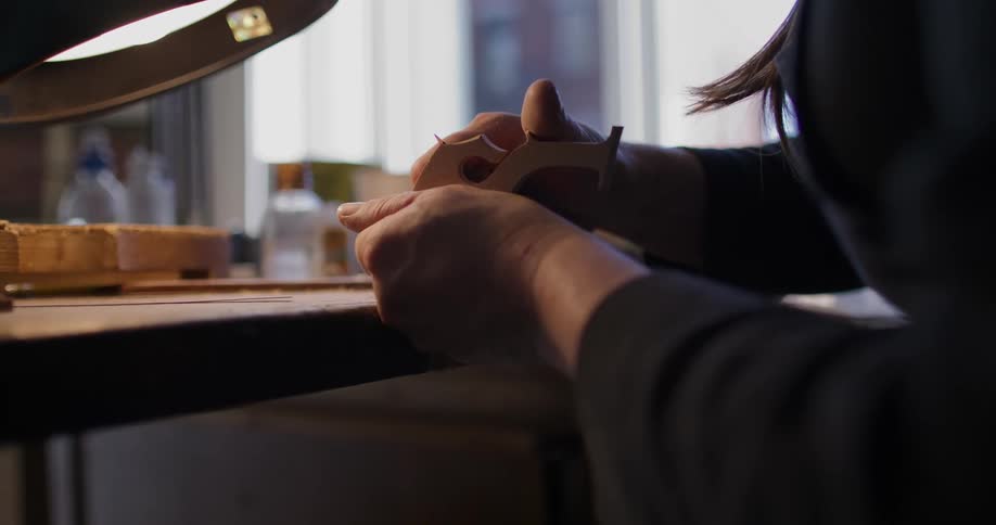 Woman carves a wooden design with a small knife at a table in front of the window