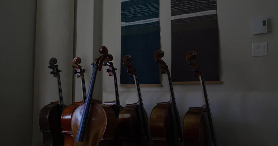 Seven cellos in a dark room are bathed in light as a door opens