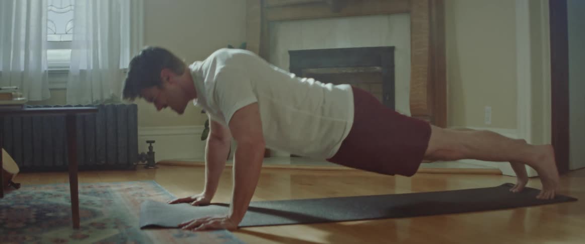 Man with a beard a white shirt and red shorts does push ups on a pilates mat in the living room at home