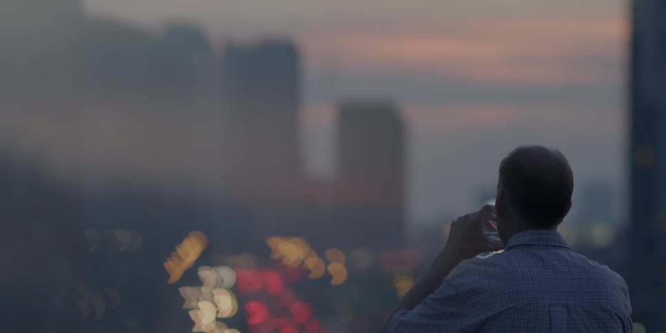 A man stands facing a city at sunset and taking a drink
