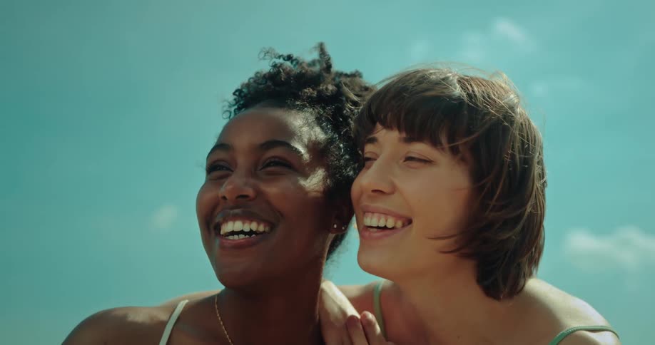 A couple wearing bikinis looks around and laughs at the beach
