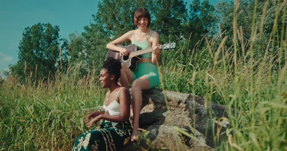 Women sitting on fallen stump among tall grasses sing and play a guitar