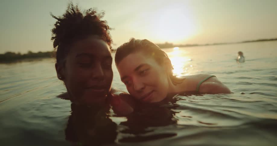 Women lean their heads together while swimming in lake as sun sits near the horizon