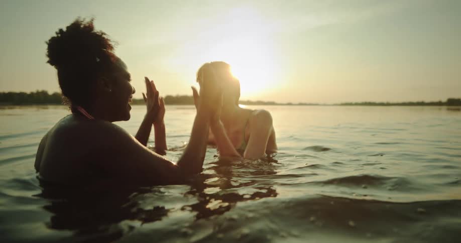 Two women inside the river clapping their hands with the sun on the horizon