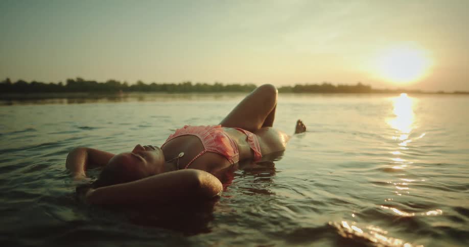 Woman wearing bikini lies in lake water reflecting sun shining from red sky